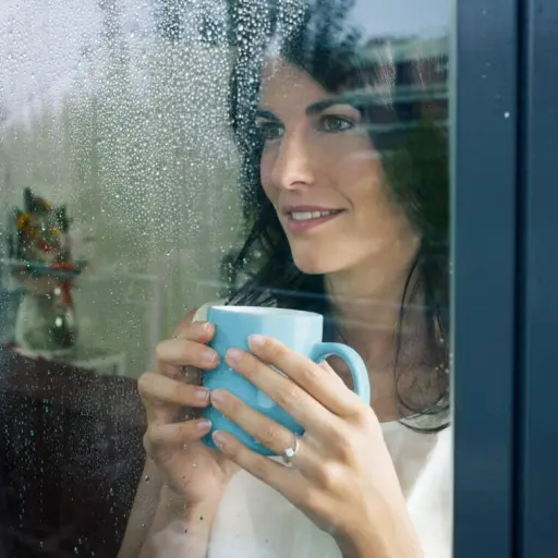 Close-up photo of a lady drinking from a mug as she looks through a glass door