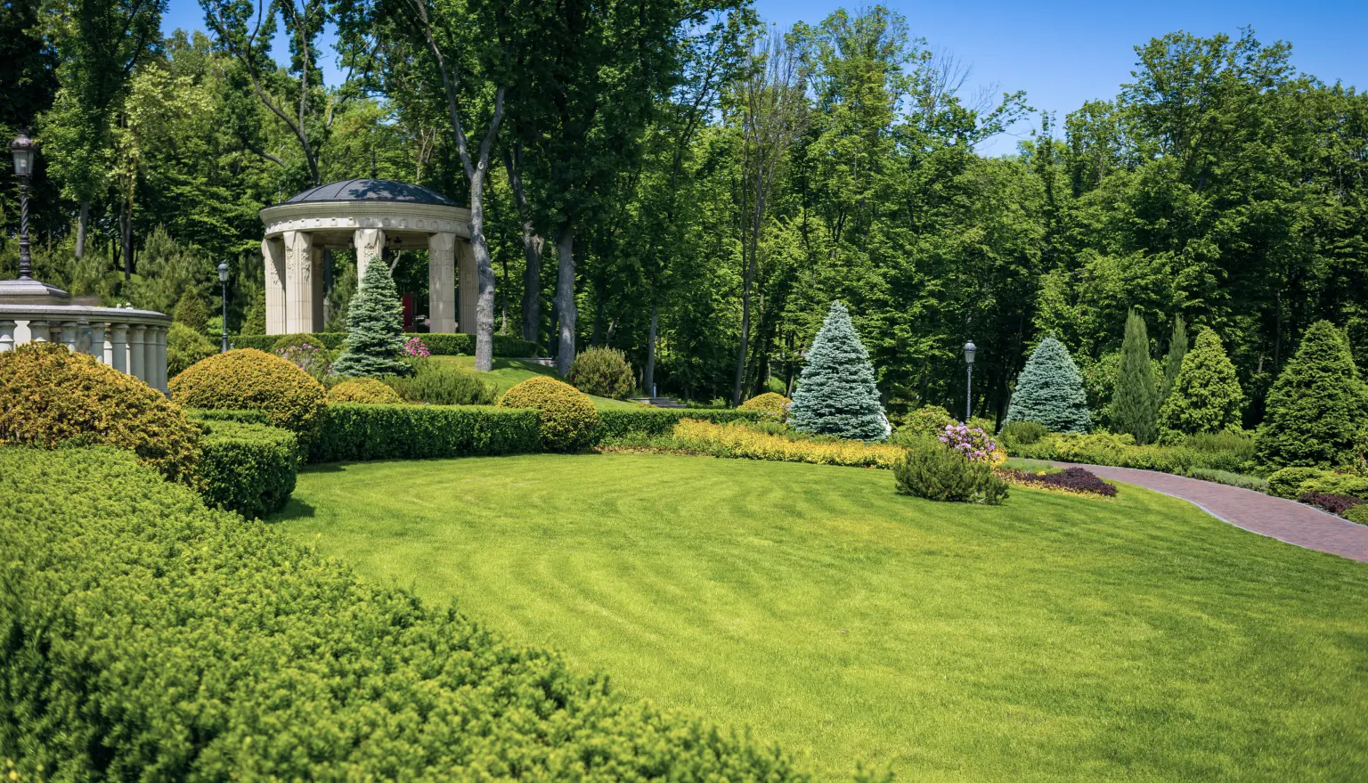 Photo of a beautiful green garden with bushes, trees, flowers, a small lamp, a small white marble building and a footpath