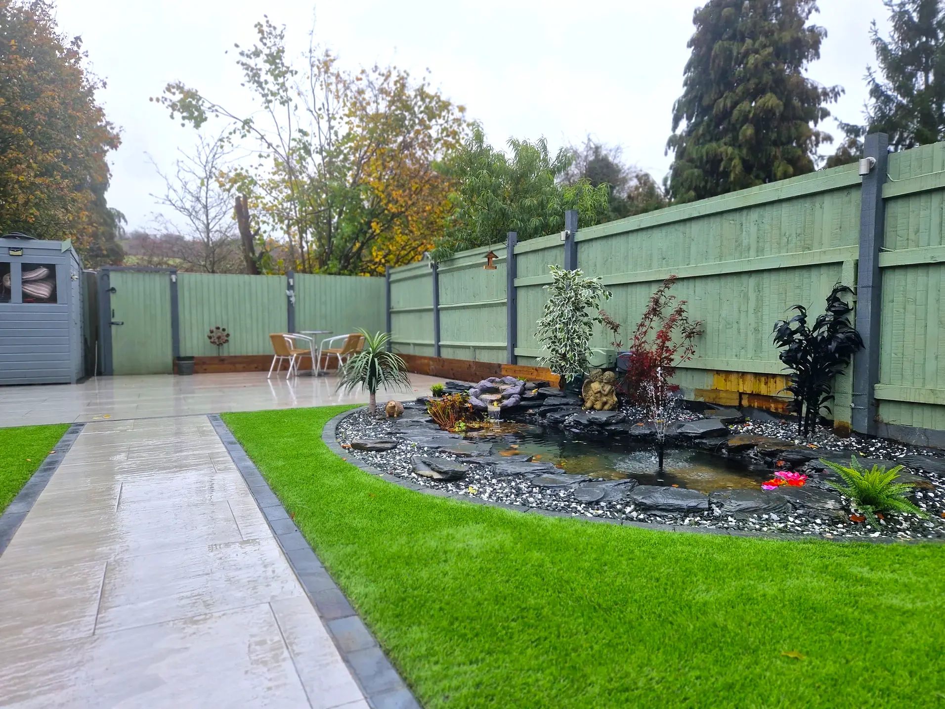 Photo of a stone patioed garden with green grass and a black stone pond with plants