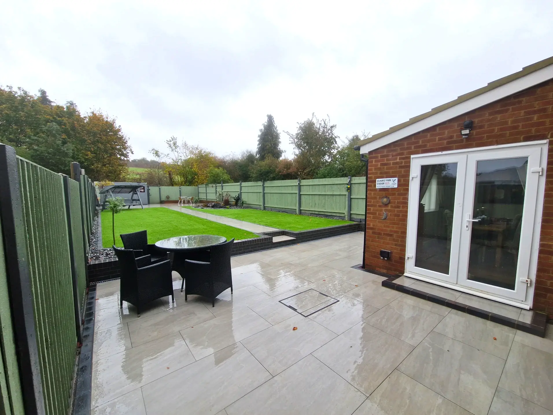 Wider photo of a garden with stone patio, green grass and a black stone pond with plants